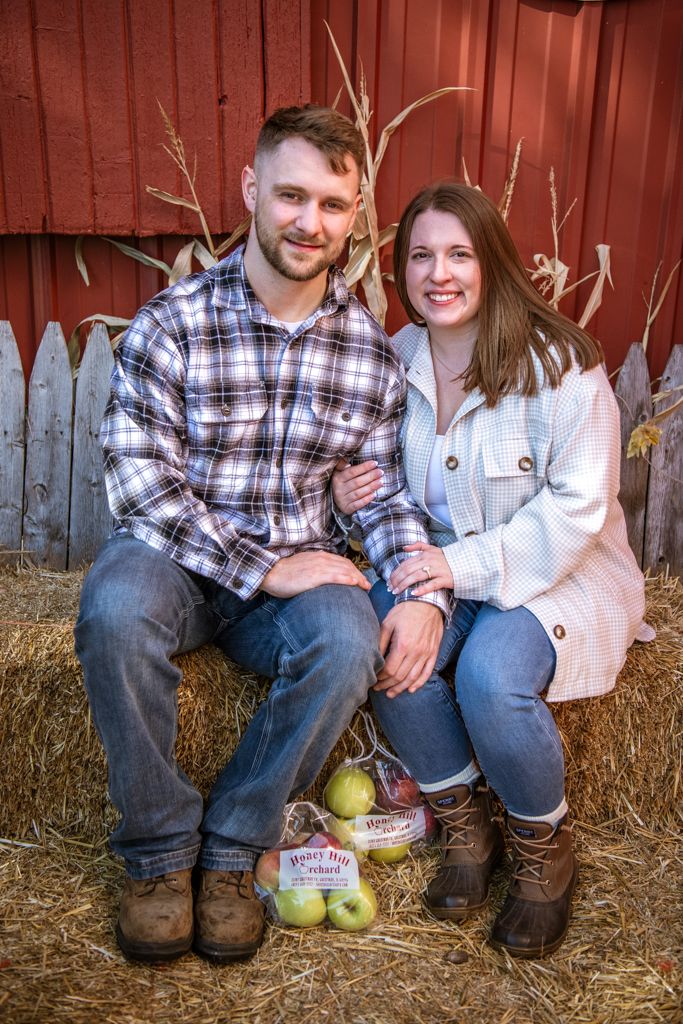 Couple sits on hay bale by red barn with apples and fall decor.