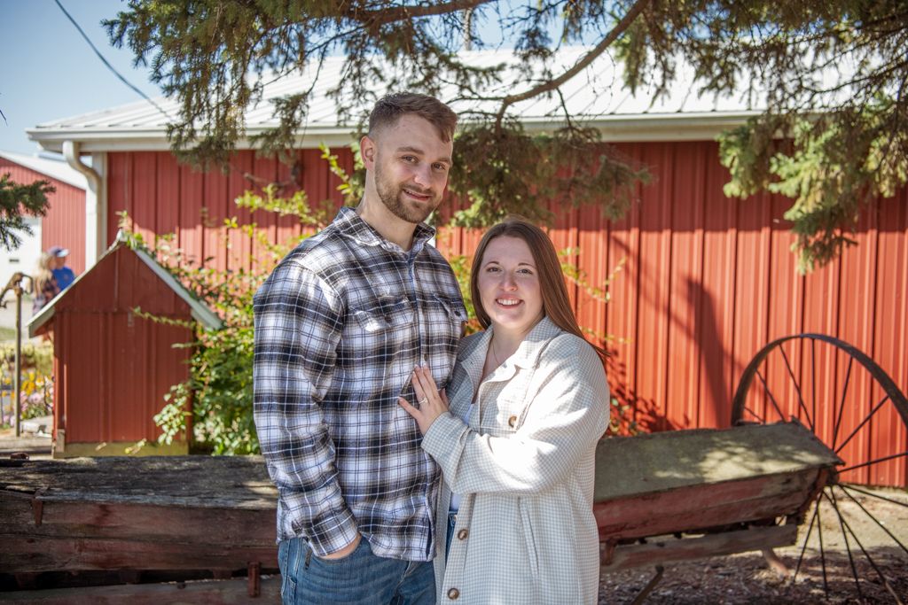 Couple poses in front of red barn wearing casual plaid and cream colored outfits.