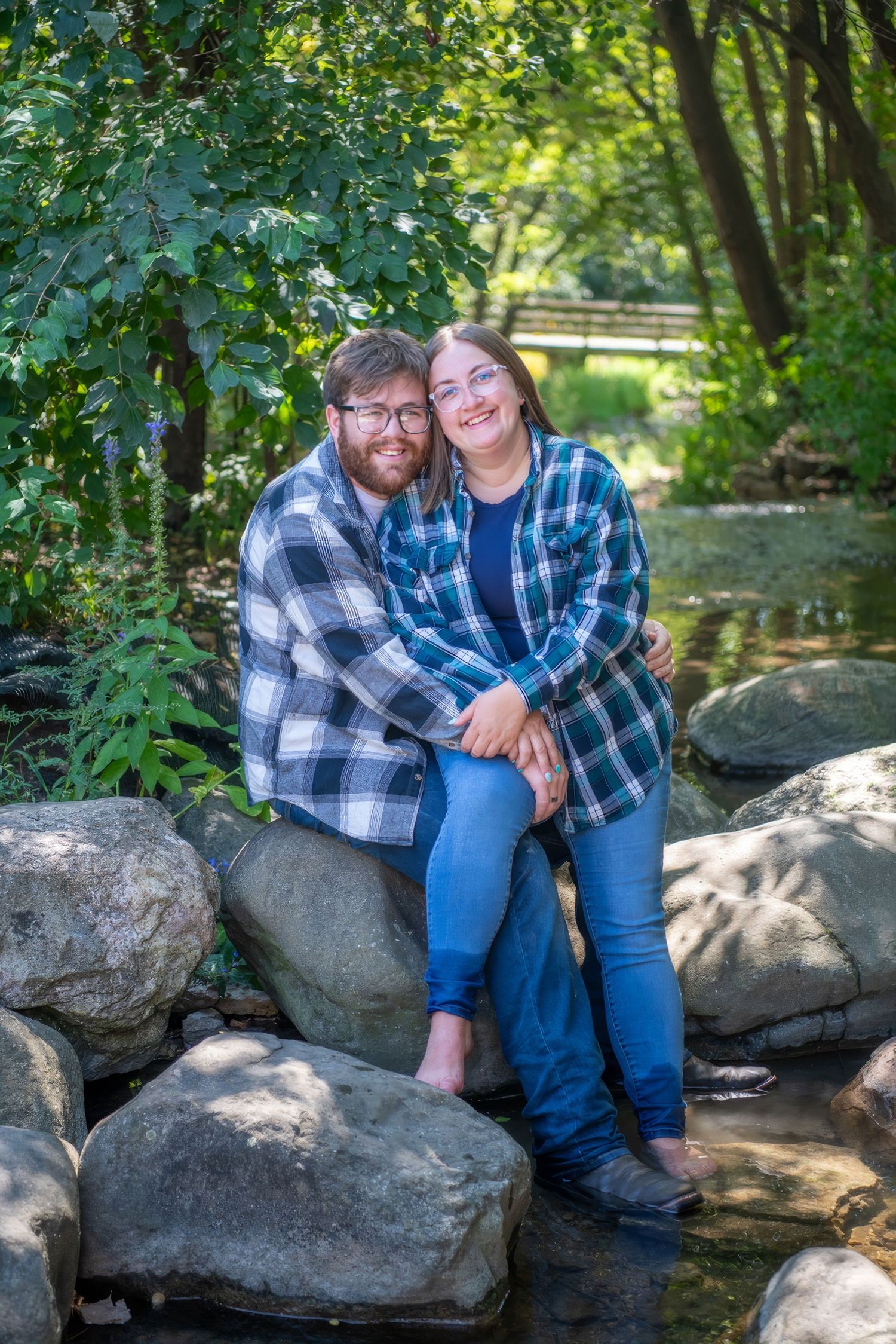 Couple wearing matching plaid shirts sitting together on large rocks by a stream.