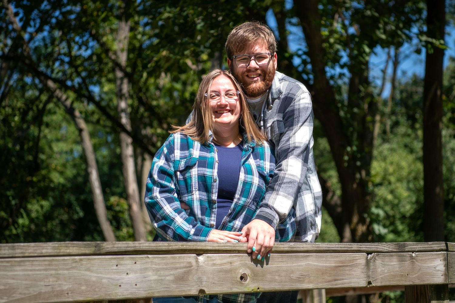 A couple leaning against a wooden fence in a sunlit outdoor setting.