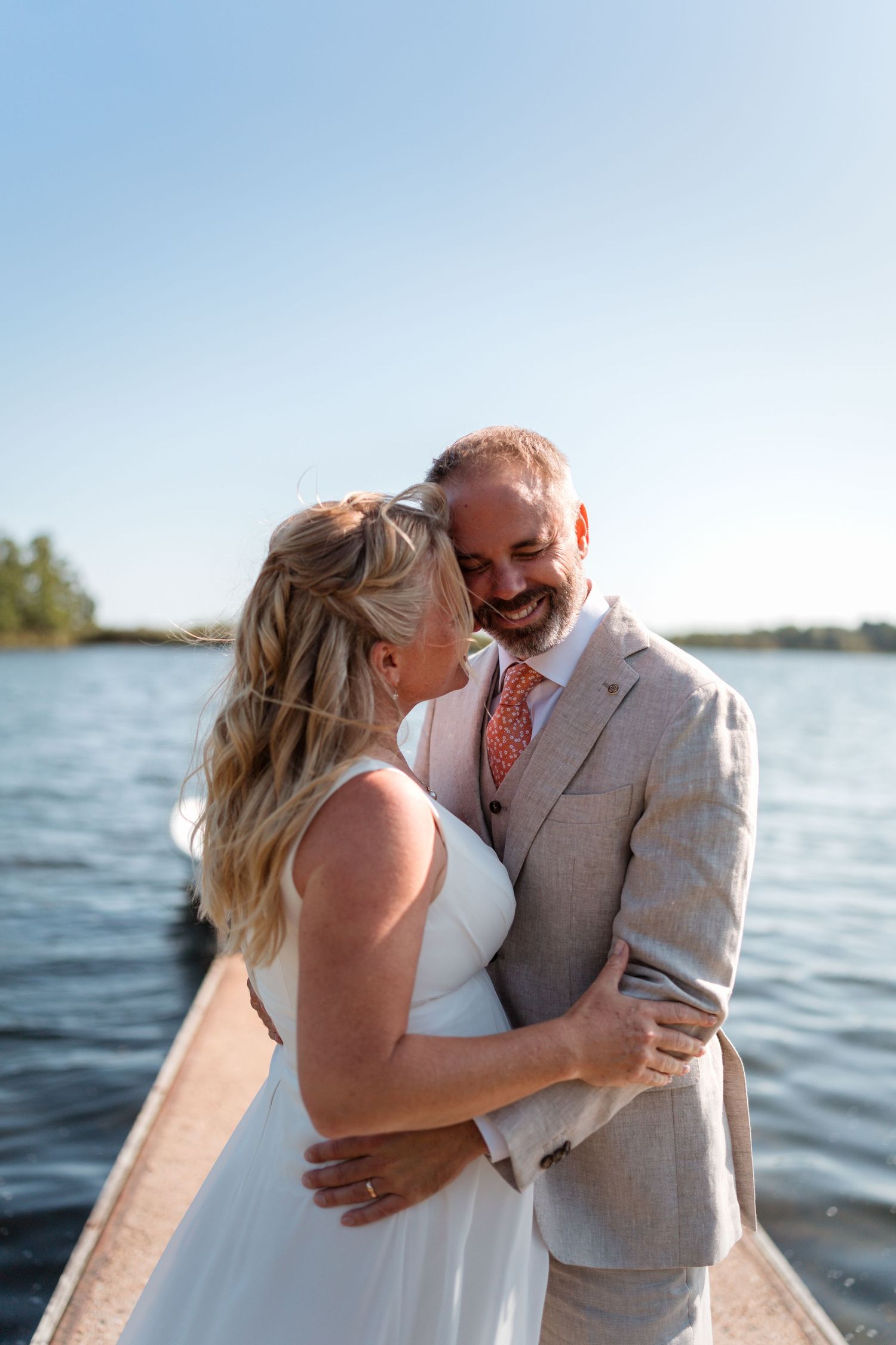 A romantic waterside wedding photo showing a bride in white and groom in black embracing by sparkling water.