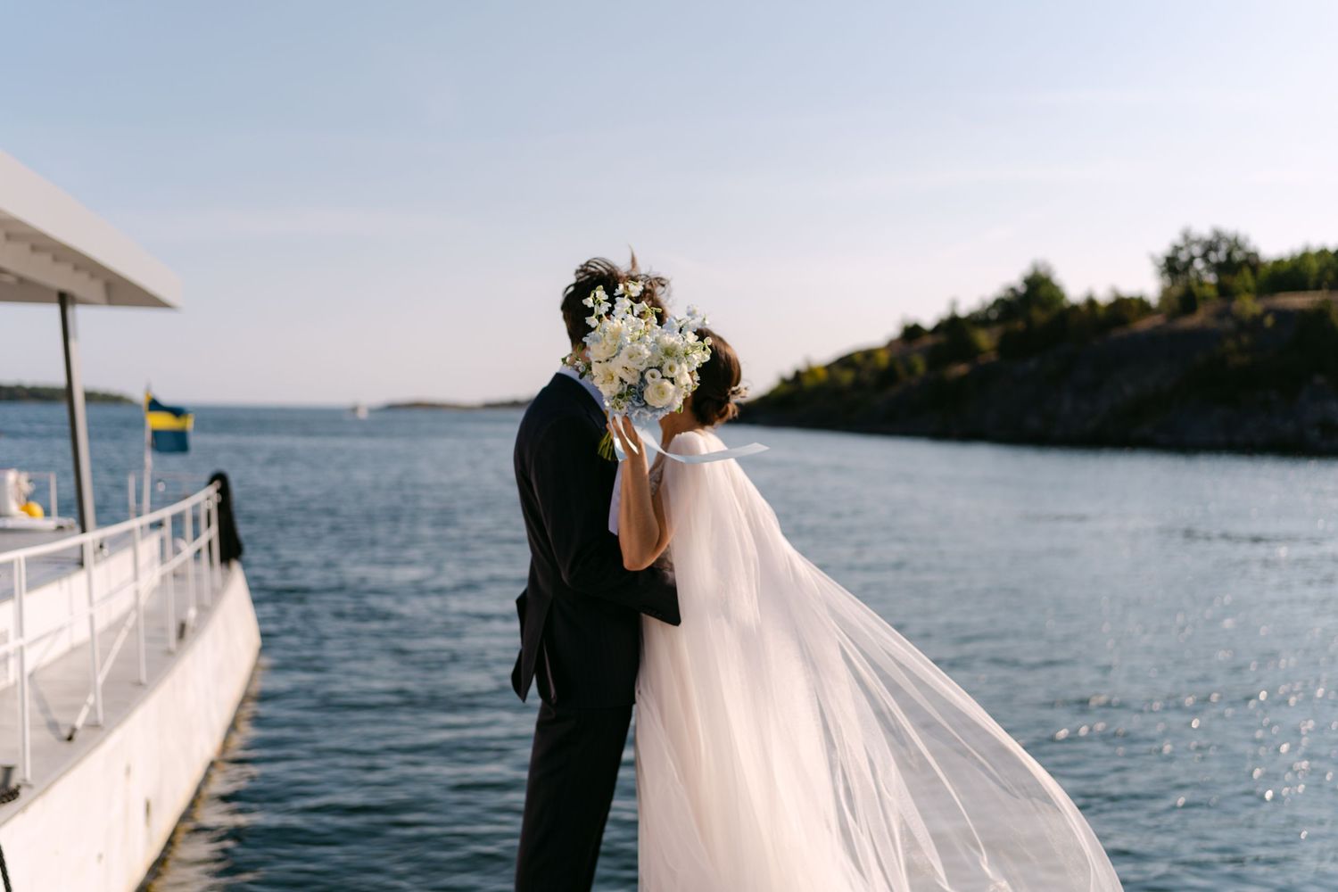 A romantic waterside wedding photo showing a bride in white and groom in black embracing by sparkling water.