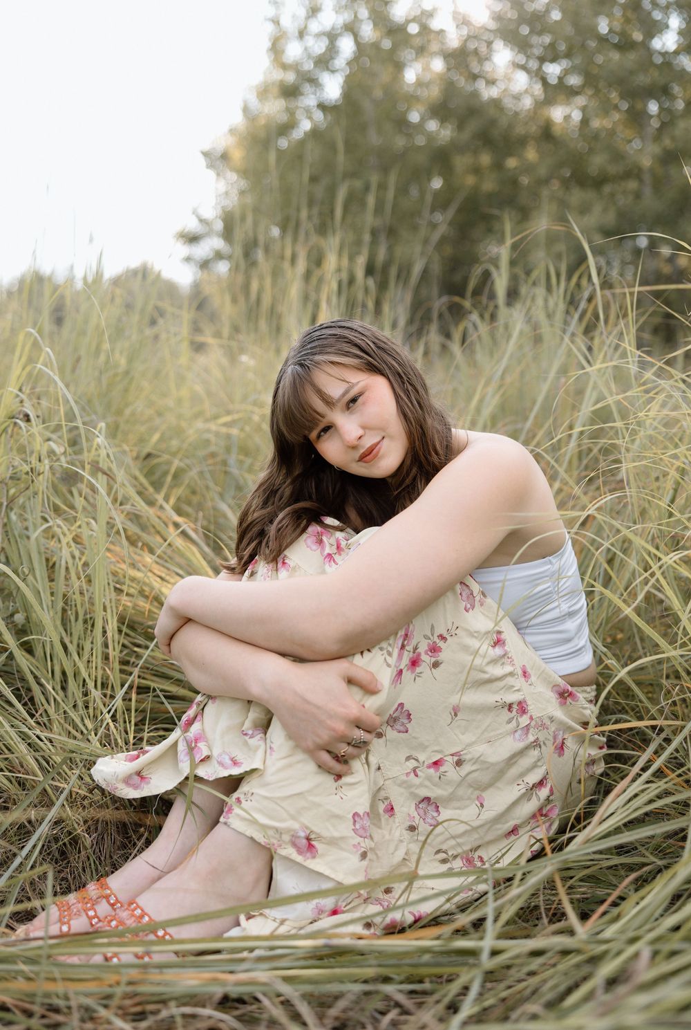 Someone in a white top and floral dress poses gracefully in tall grass during sunset.