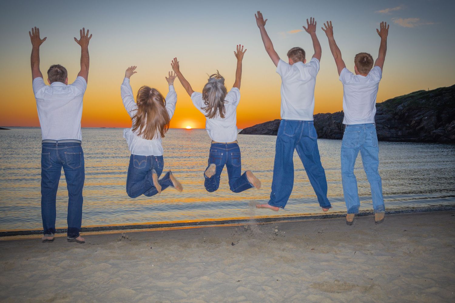 Family of five jumping joyfully on a beach at sunset with arms raised.