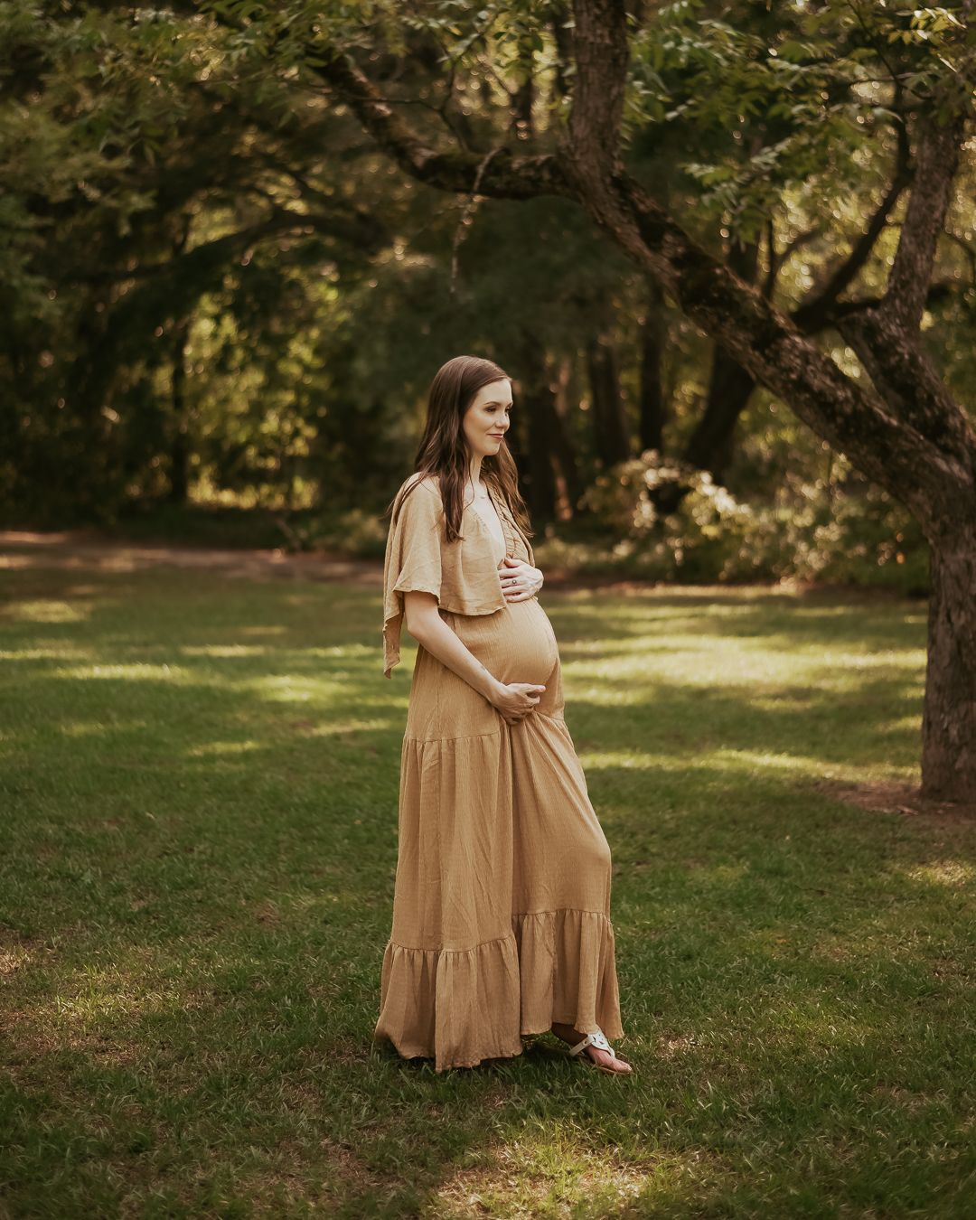 Expecting mother in flowing tan dress poses in sunlit garden setting.