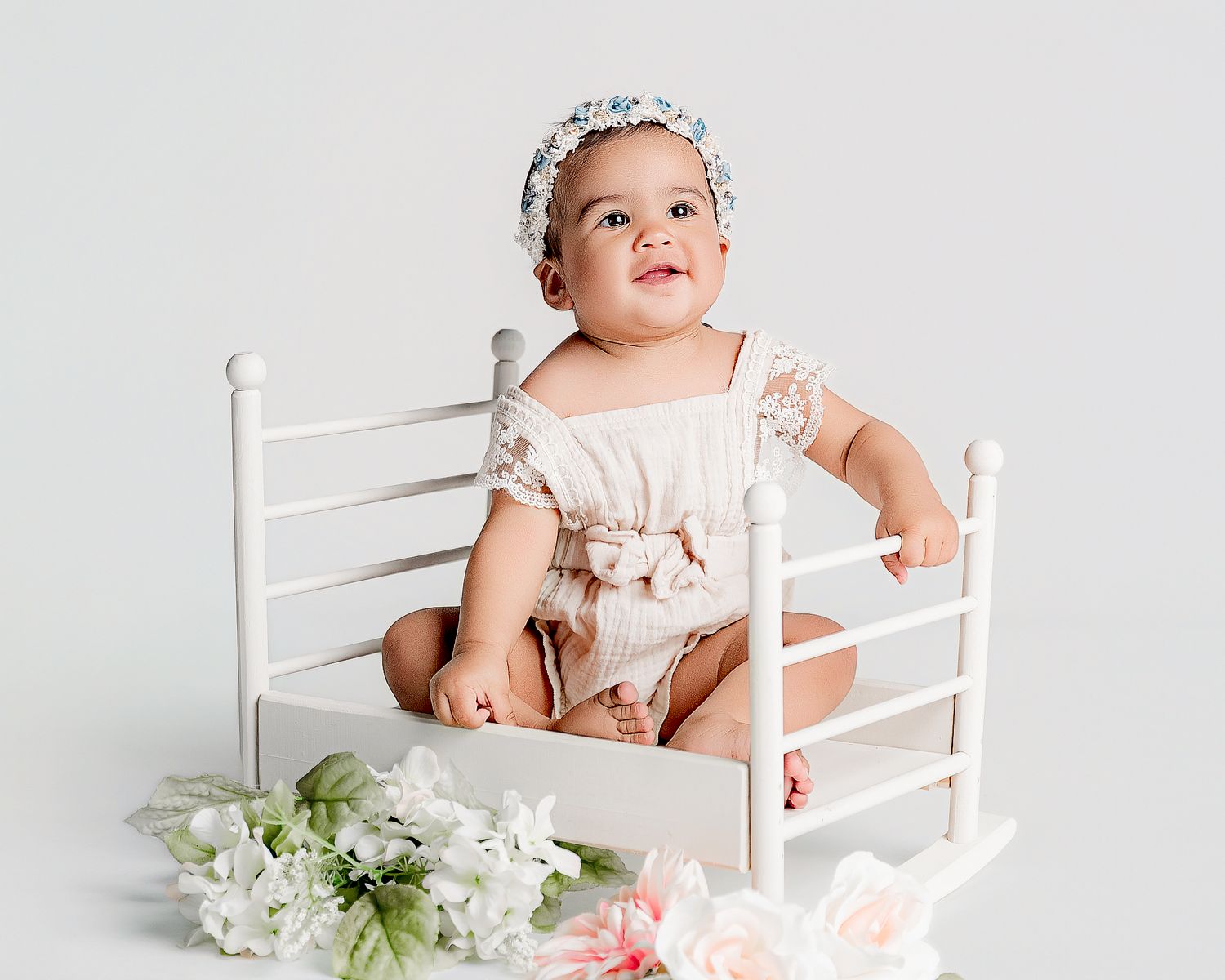 Baby in white dress sits in rustic chair with delicate floral decorations for professional portrait session.