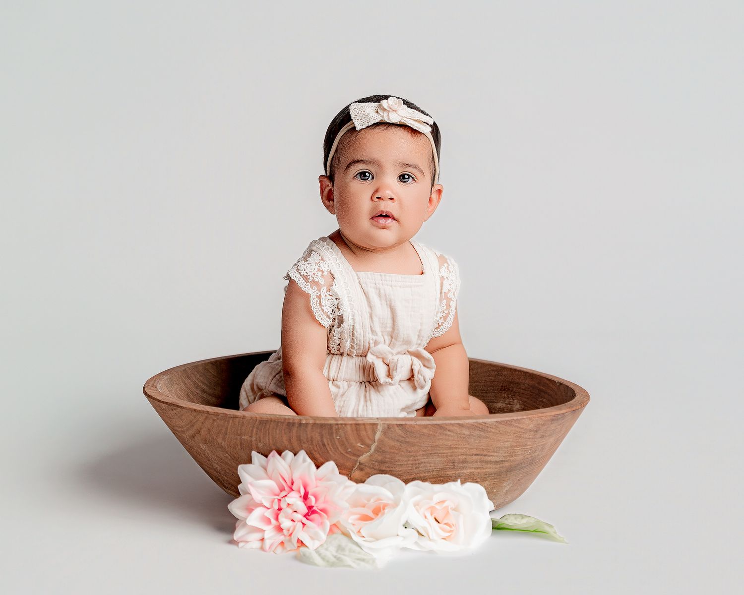 Small baby wearing white outfit sits in rustic wooden bowl with pink and peach flowers displayed in front.