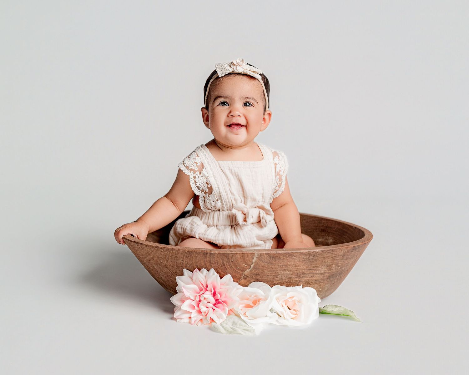 A baby wearing a white dress sits in a wooden bowl with pink flowers against a light gray background.