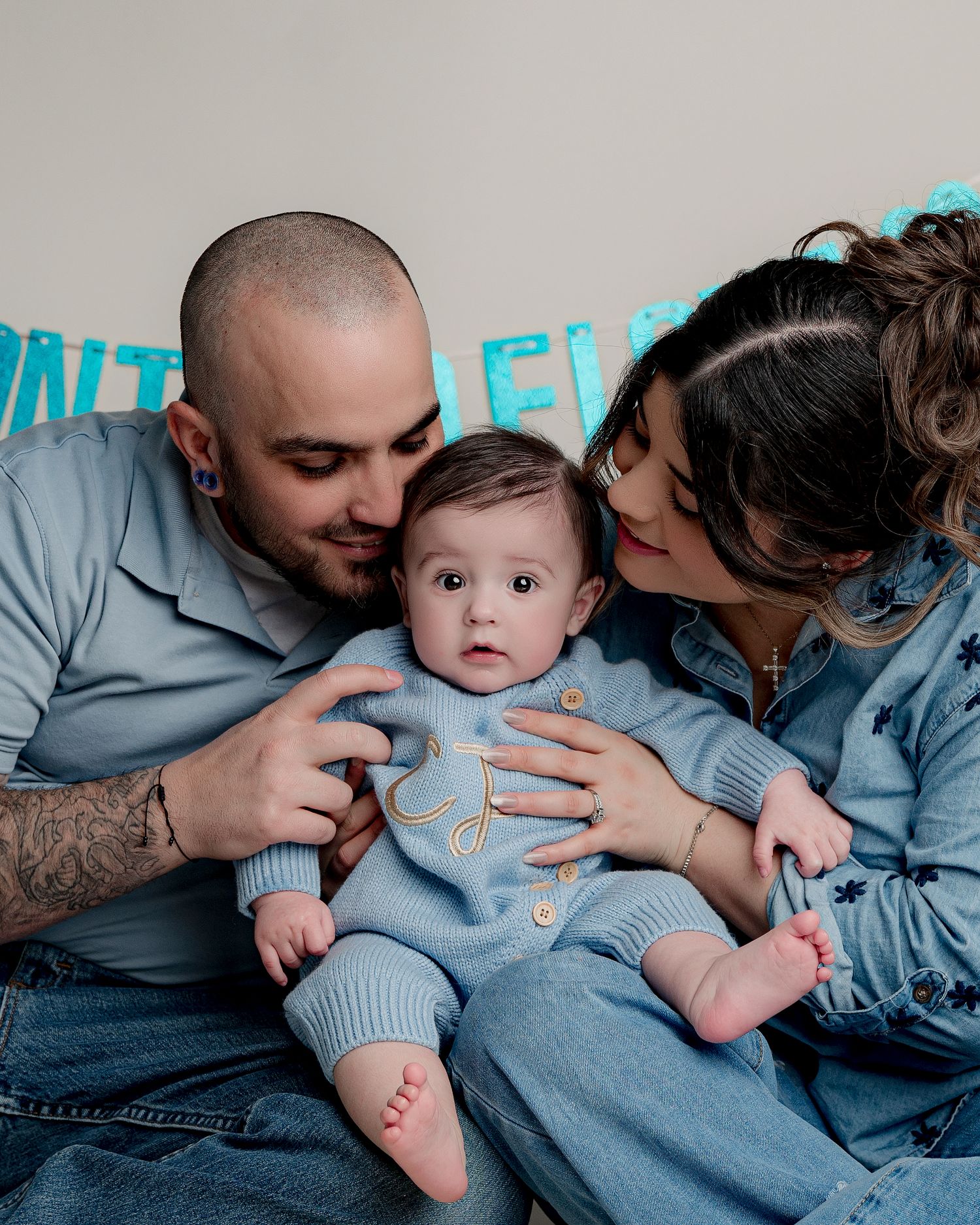 Parents in denim sharing intimate moment with baby under turquoise banner.