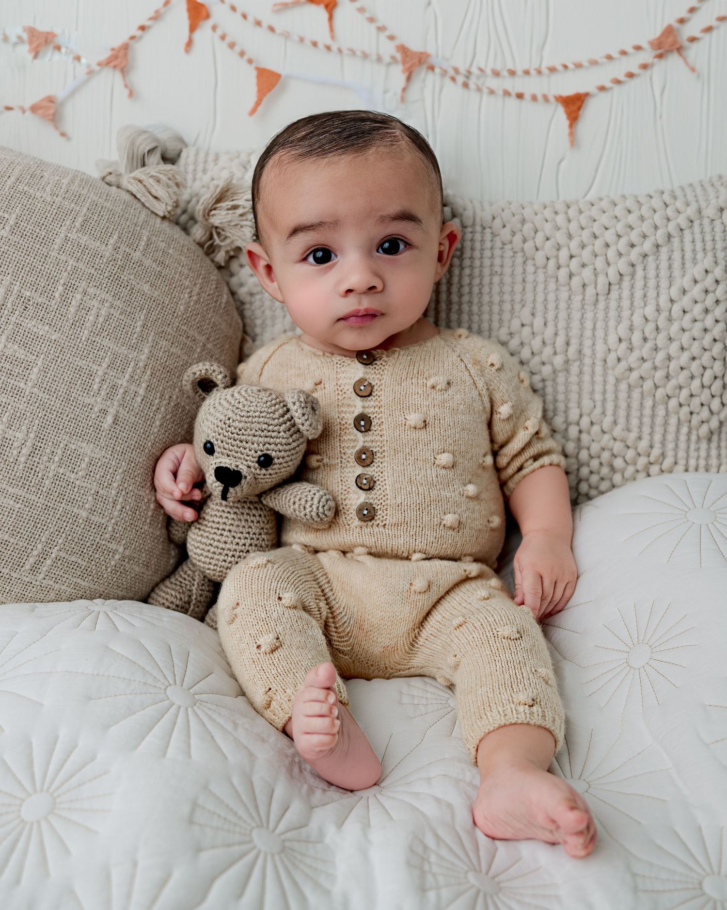 Baby in cream knit outfit sitting on couch with teddy bear and coral bunting backdrop.
