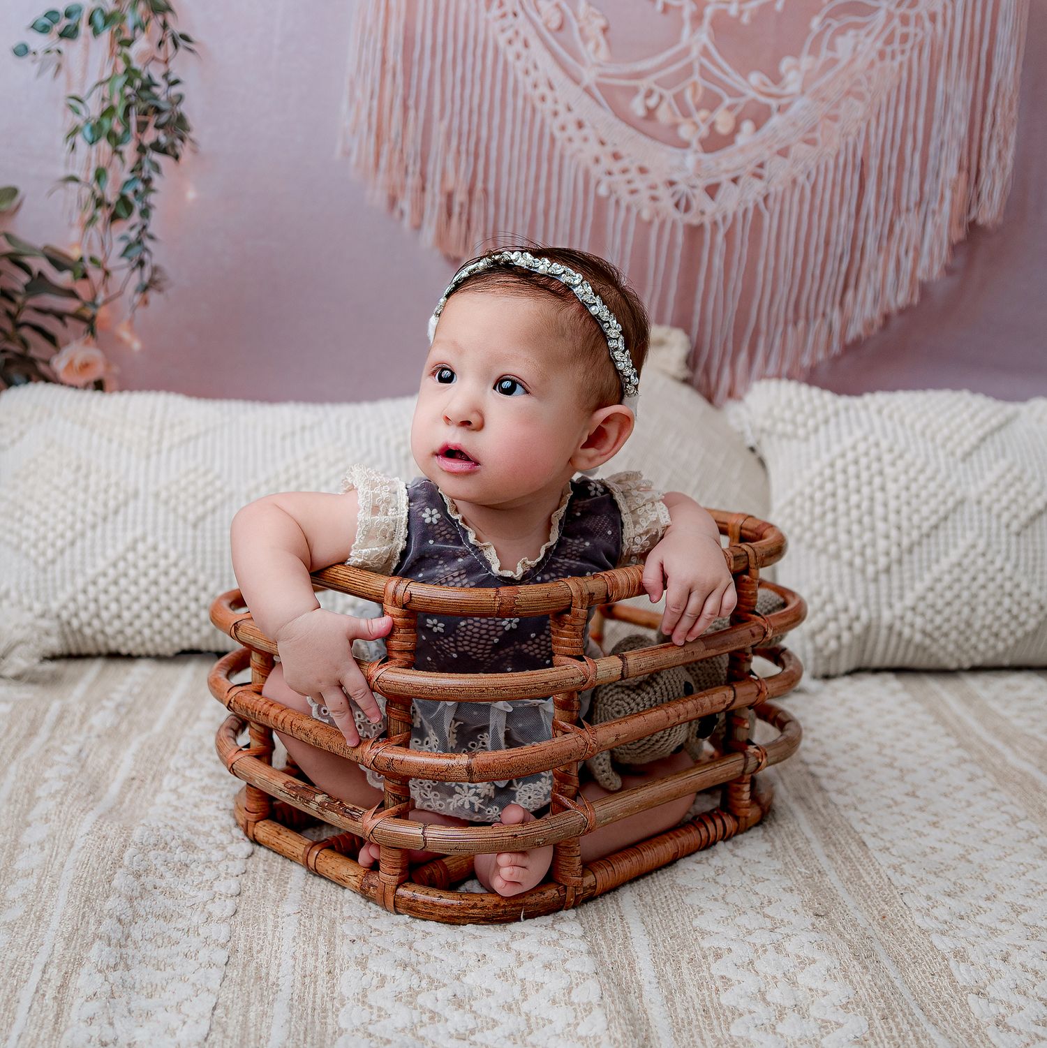 Baby wearing gray dress poses in rustic wooden basket with macrame backdrop.