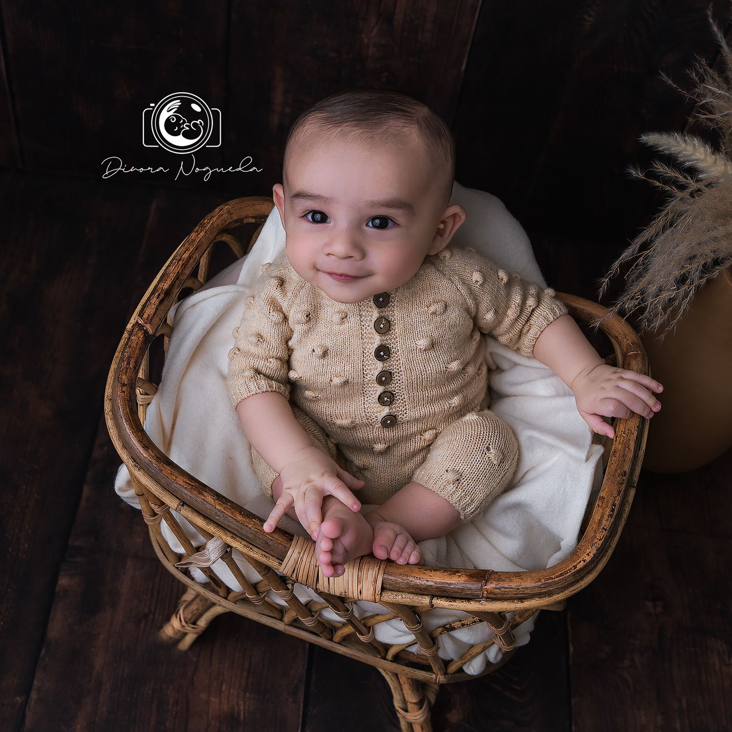 Smiling baby in beige knit romper sitting in vintage wicker chair.