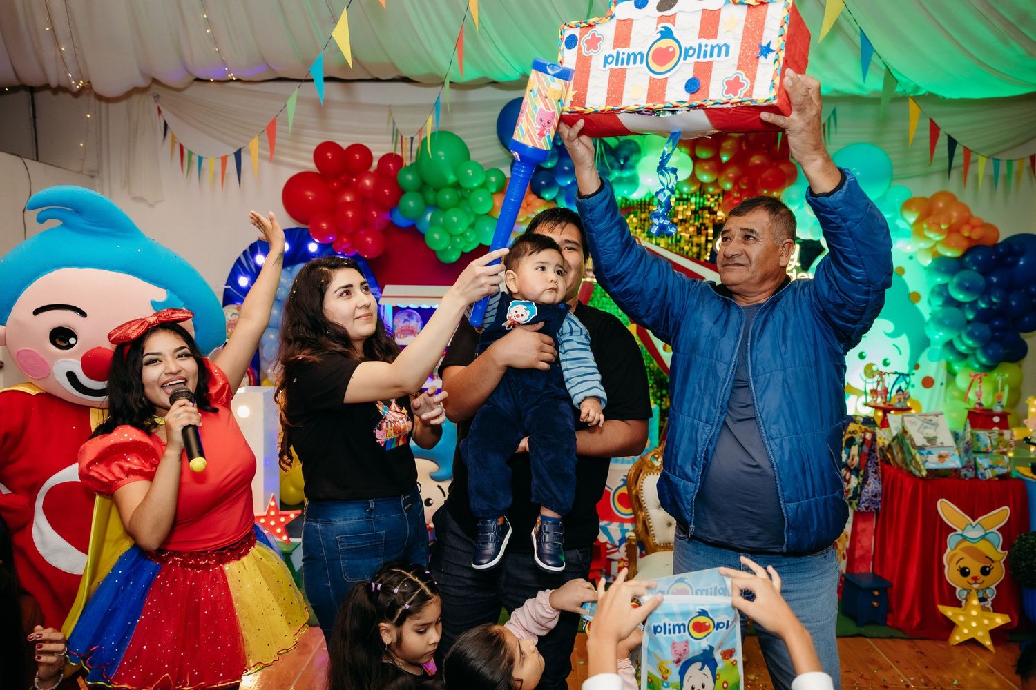 Colorful children's party with guests celebrating under festive decorations and balloons.