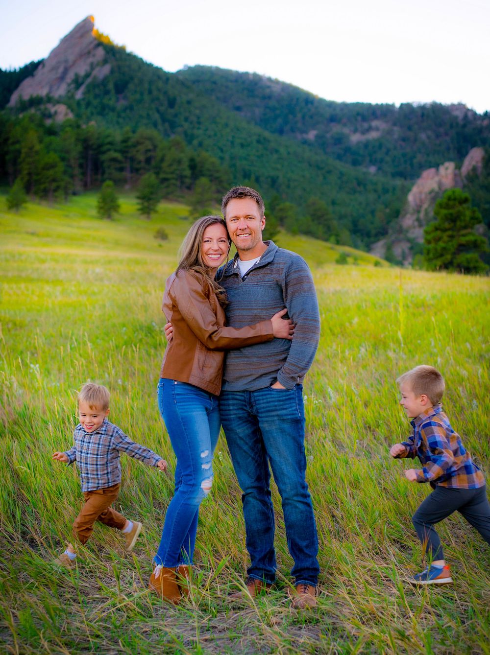 Family photo in mountain meadow with playful children running through tall grass.