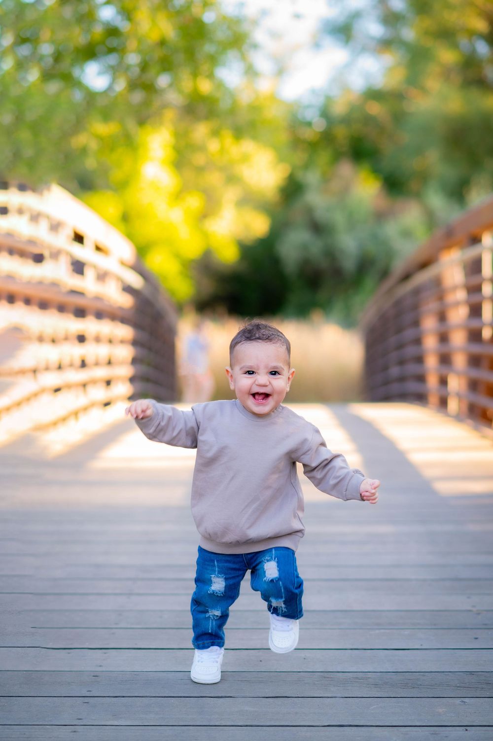 Toddler in gray sweater and jeans taking first steps on wooden bridge walkway.