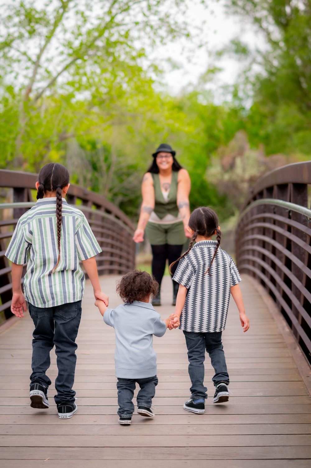 Three children walk hand-in-hand on a wooden bridge toward a smiling woman in a park setting.