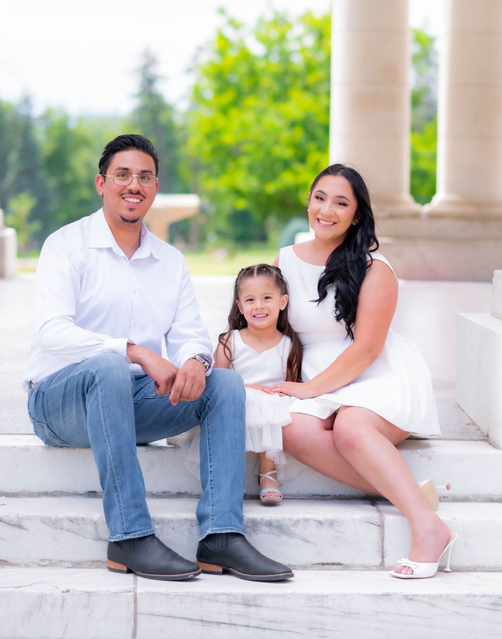 A happy family of three in white outfits sitting on stone steps outdoors, with lush green trees in the background.