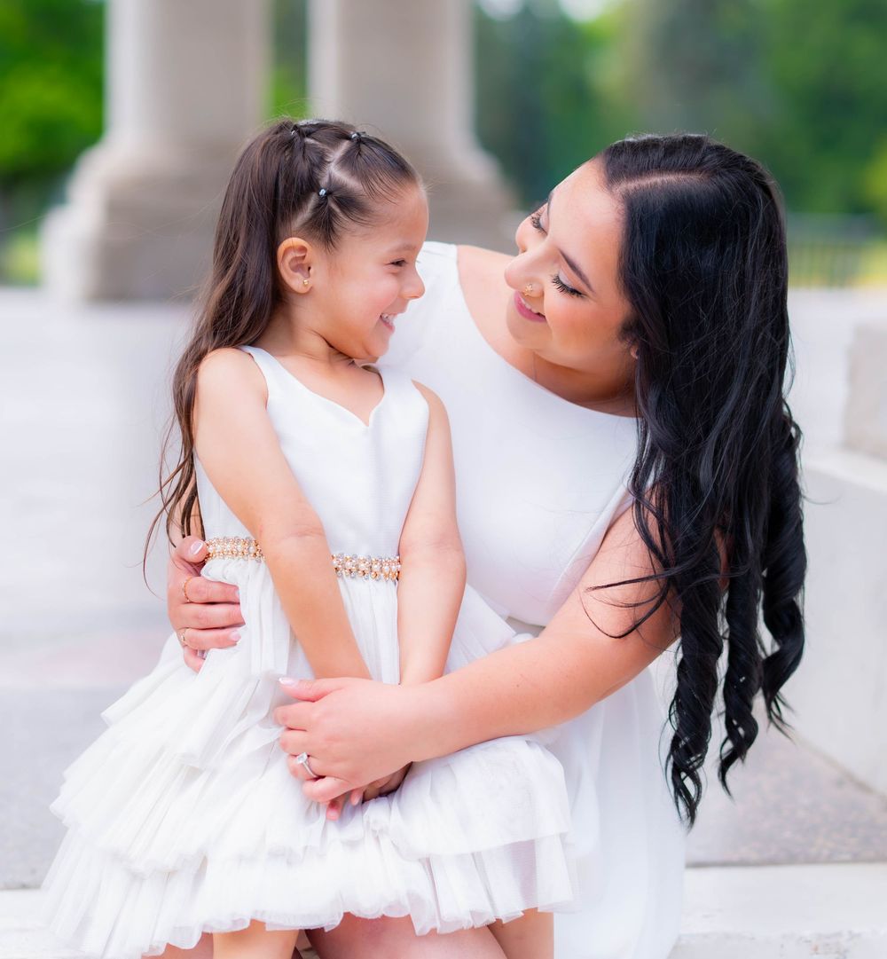 A woman in a white dress lovingly embraces a young girl in a white ruffled dress, sharing a joyful moment outdoors.