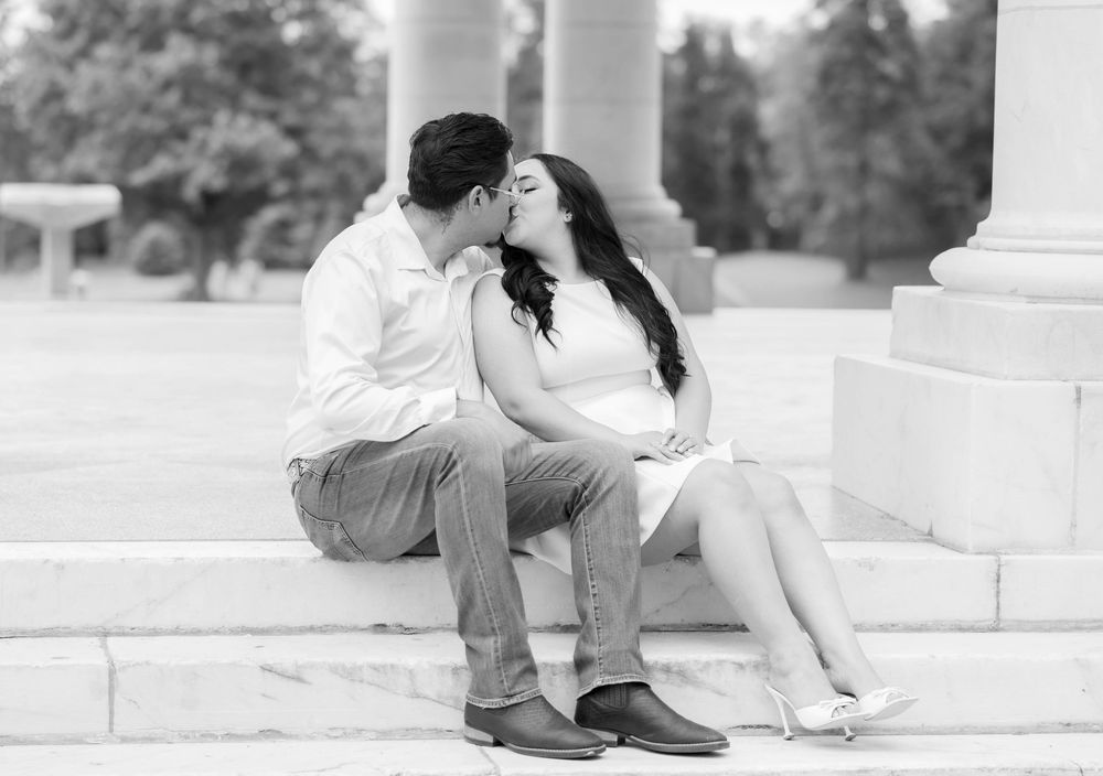 Black and white photo of a couple sharing a romantic kiss while sitting on marble steps outdoors.