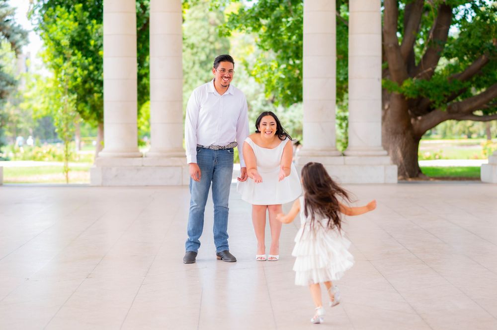 Family of three enjoying a sunny day outdoors, with a little girl running toward her parents near elegant white columns.
