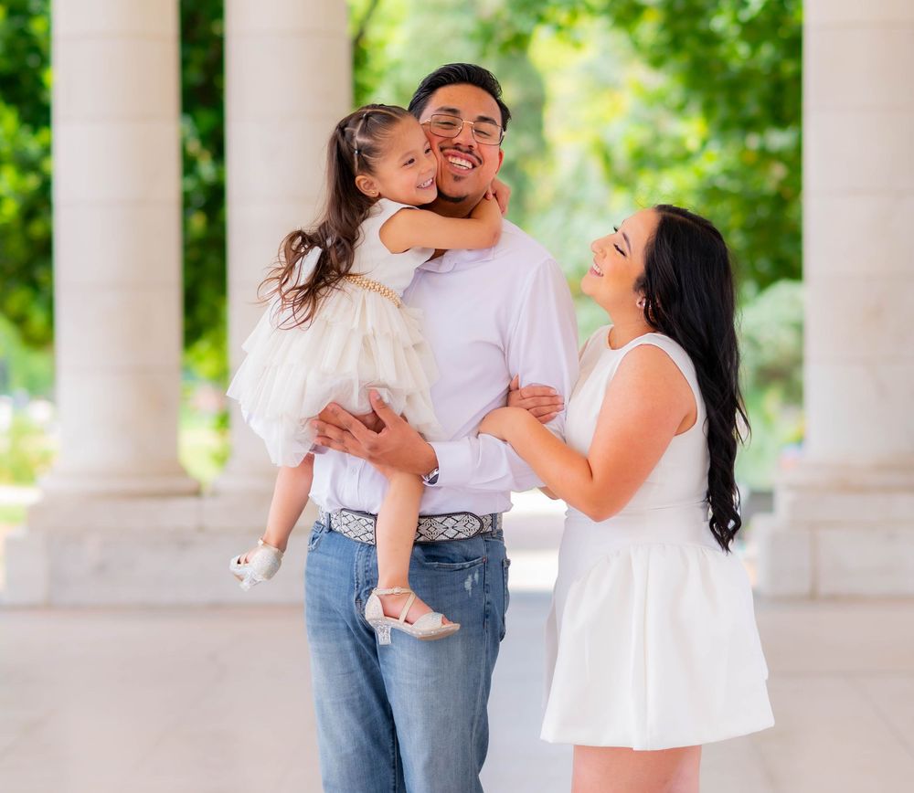 A joyful family portrait with a man holding a young girl who kisses his cheek, beside a smiling woman in white.
