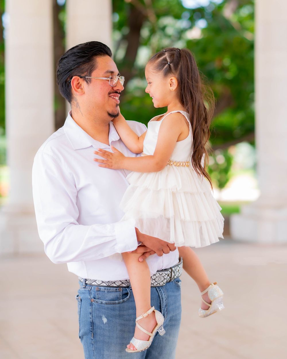 A father holds his young daughter in a white dress outdoors, sharing a tender moment near white columns.