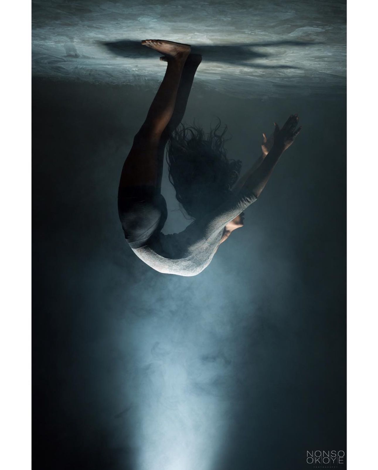 Artistic underwater photograph of figure diving in dark blue waters with dramatic lighting.