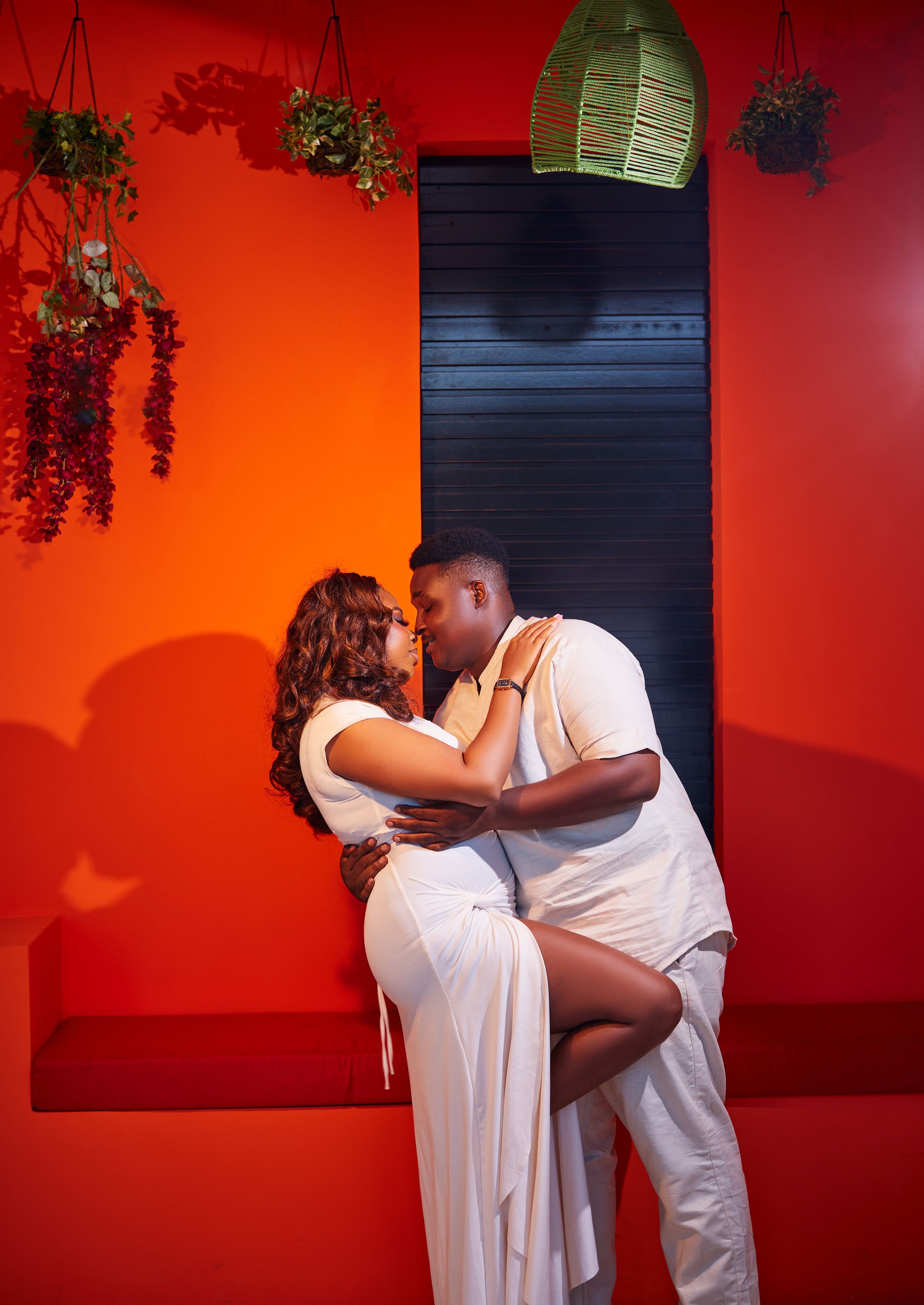 Romantic couple embracing against vibrant orange wall with hanging plants.