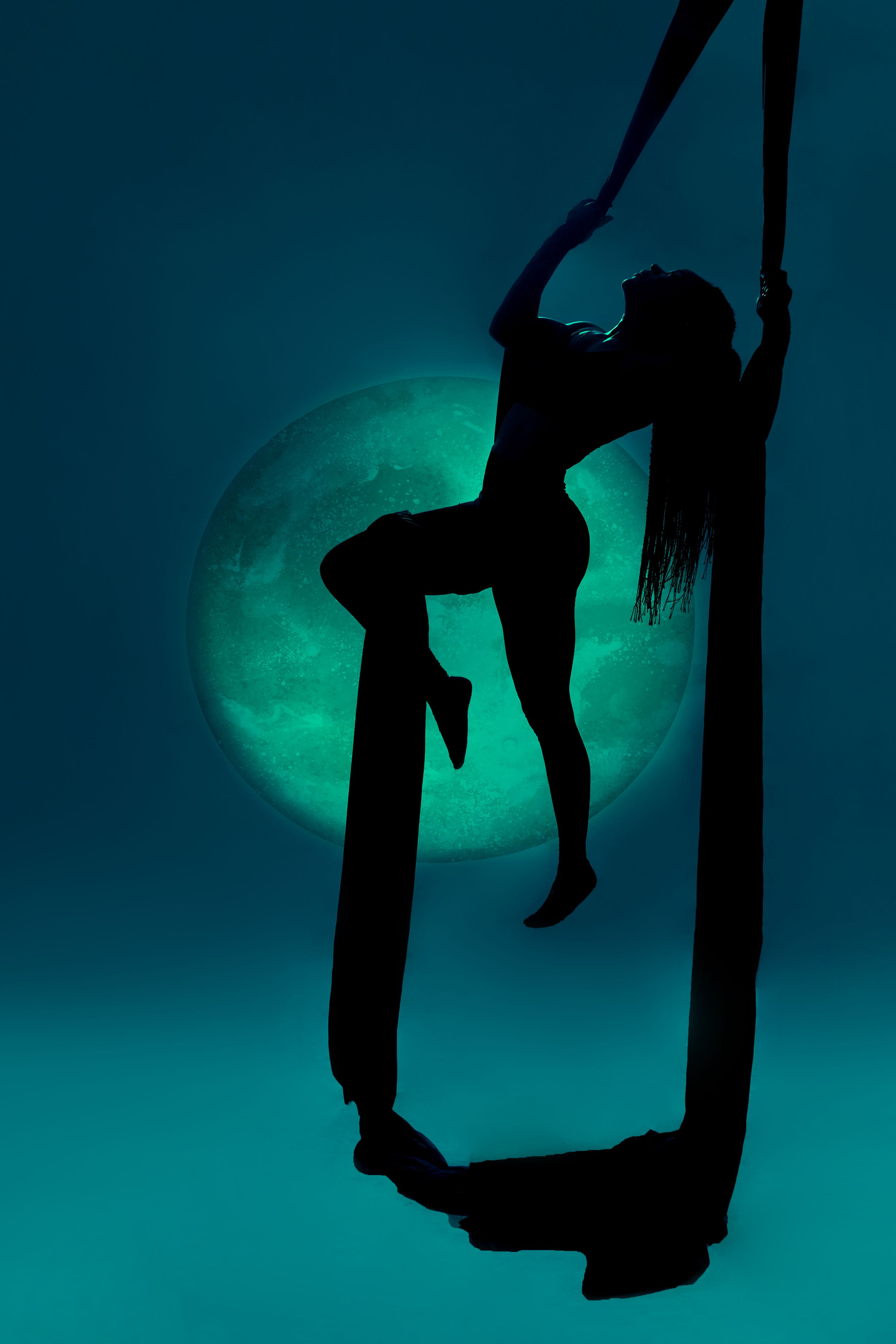 Silhouette of an aerial performer against a glowing green moon in a dark turquoise sky.