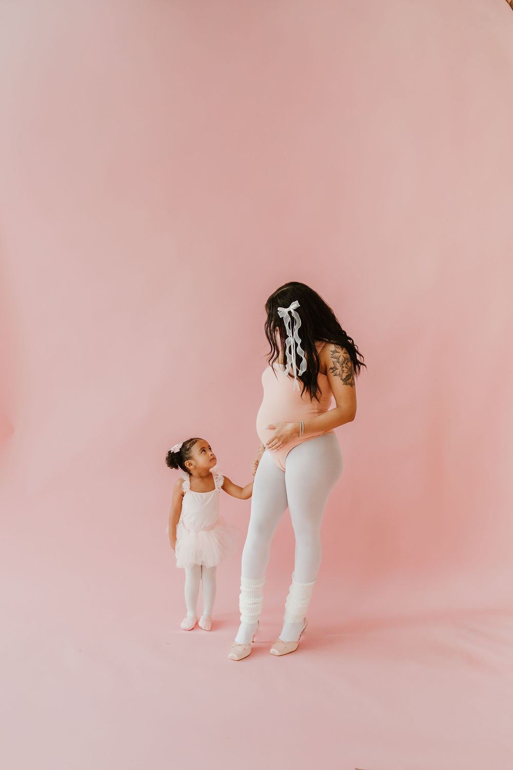 Mother and daughter in matching white outfits hold hands against a pink backdrop during a minimalist photo session.