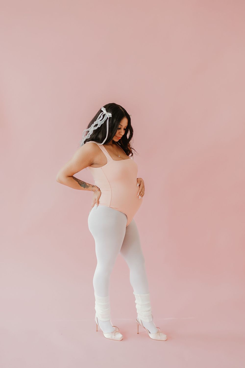 Expectant mother in white outfit poses against pink backdrop during maternity photoshoot.