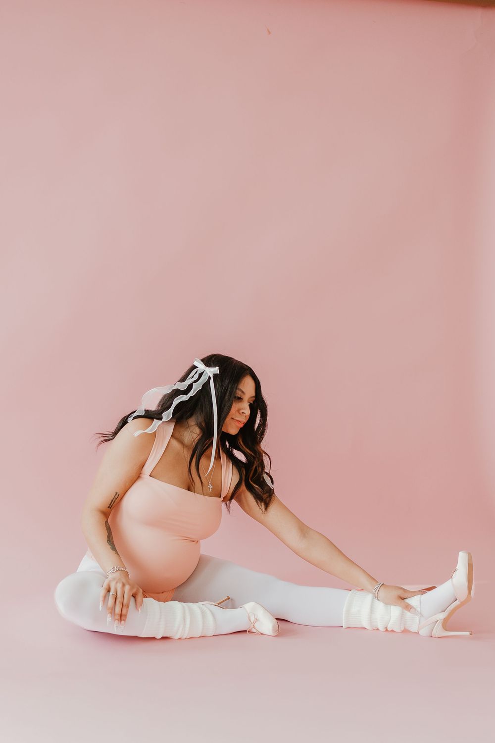 A pregnant woman in a pink bodysuit and white boots stretches on a pink backdrop during a maternity photoshoot.
