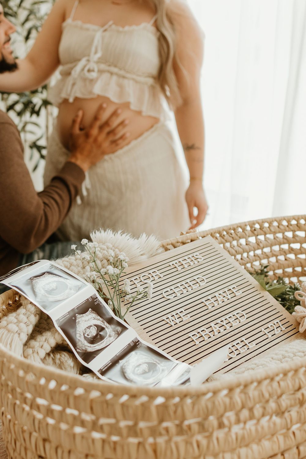 Woven basket holds ultrasound photos and baby's breath flowers while a pregnant woman in white dress stands nearby.