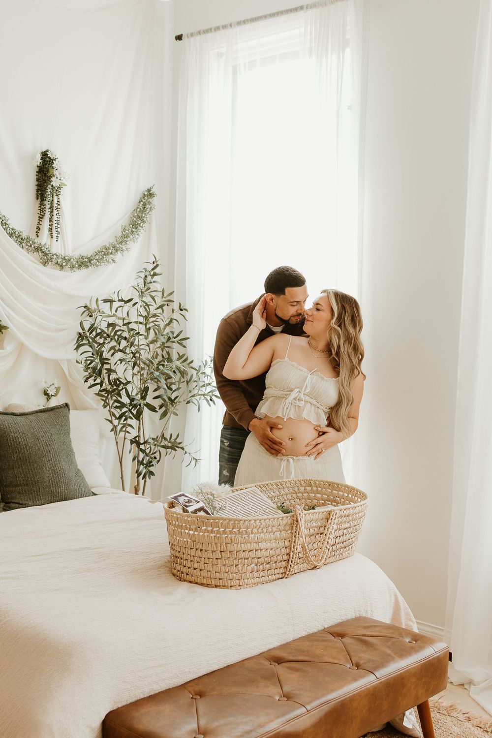 Intimate maternity photoshoot in bright white room with woven basket and flowing curtains.