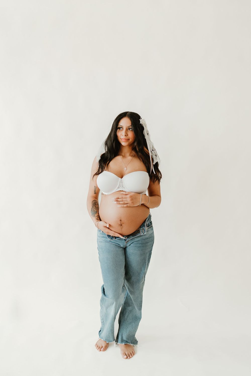 Pregnant woman in white bralette and light blue jeans poses barefoot against white backdrop for maternity photo.