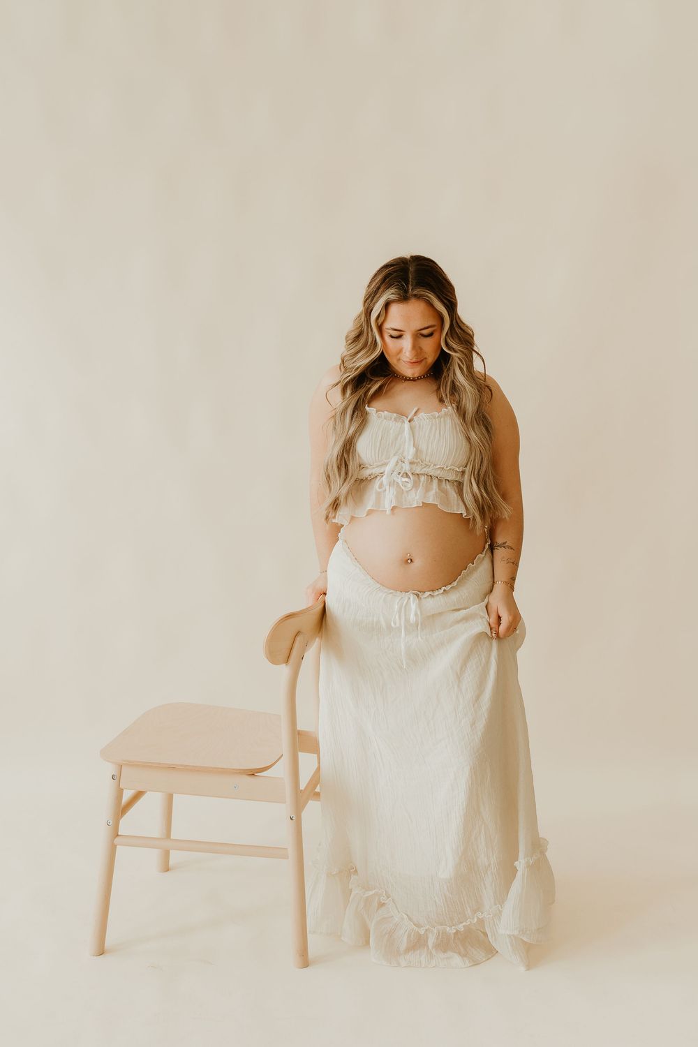 A minimalist maternity photo series showing a figure in white against a neutral backdrop posing with a wooden chair.