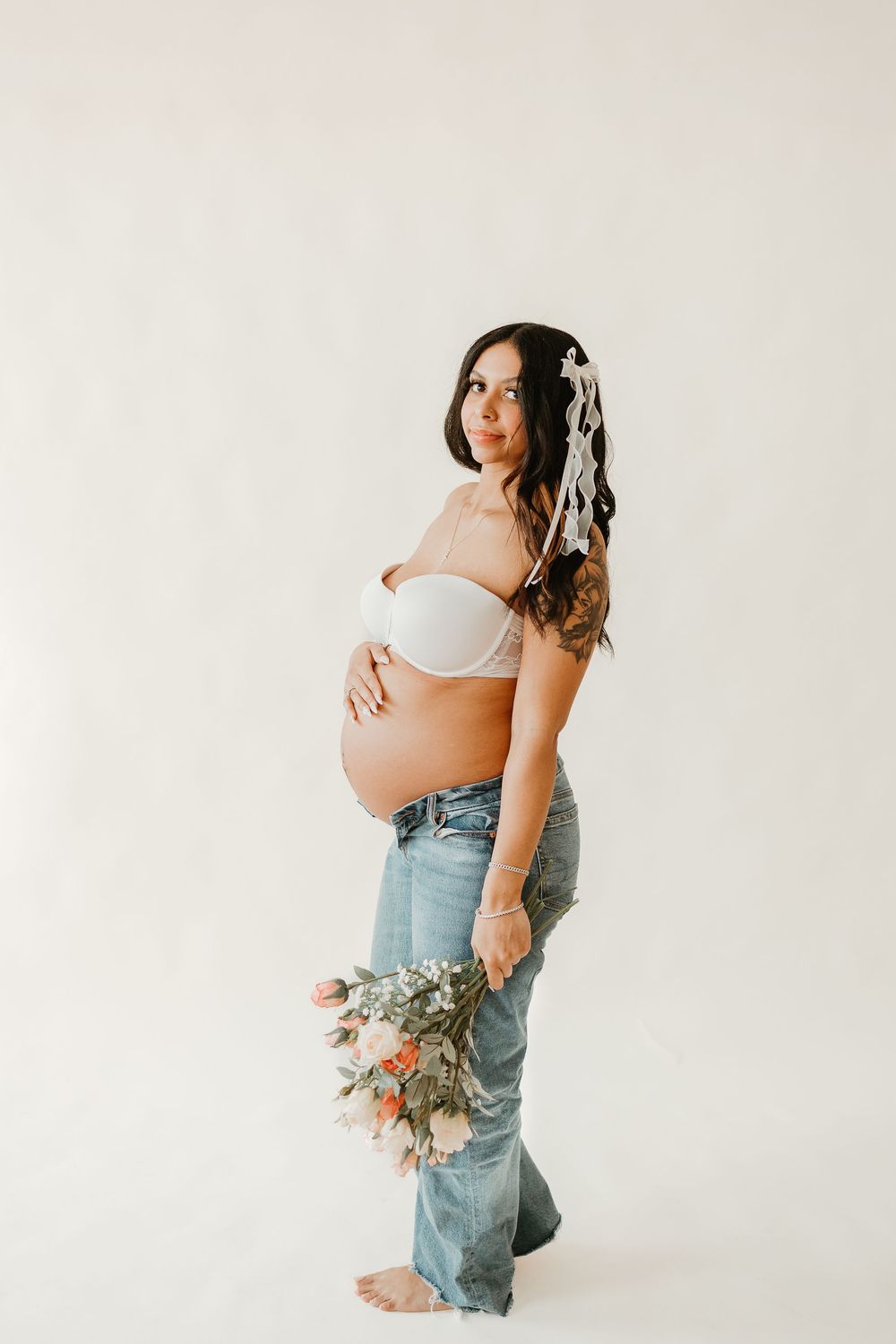 Maternity photo series showing expectant mother in white crop top and jeans against neutral background with dried flowers.