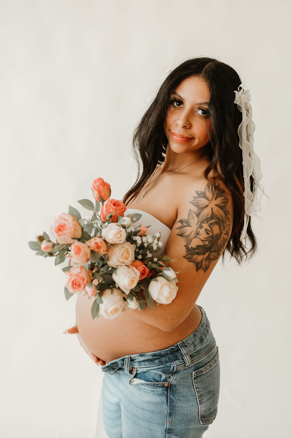 Artistic maternity photoshoot featuring a floral bouquet with peach roses and denim jeans against a white backdrop.