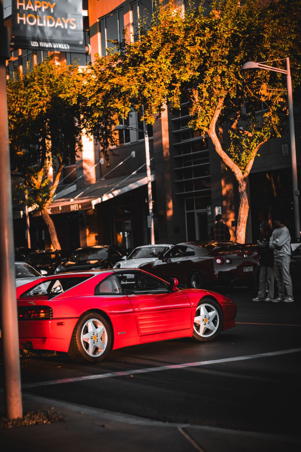 Red Ferrari parked on night street with illuminated trees and Happy Holidays sign.