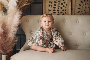 A baby girl in a floral dress and bow headband sits on a beige tufted sofa in a boho-styled room.