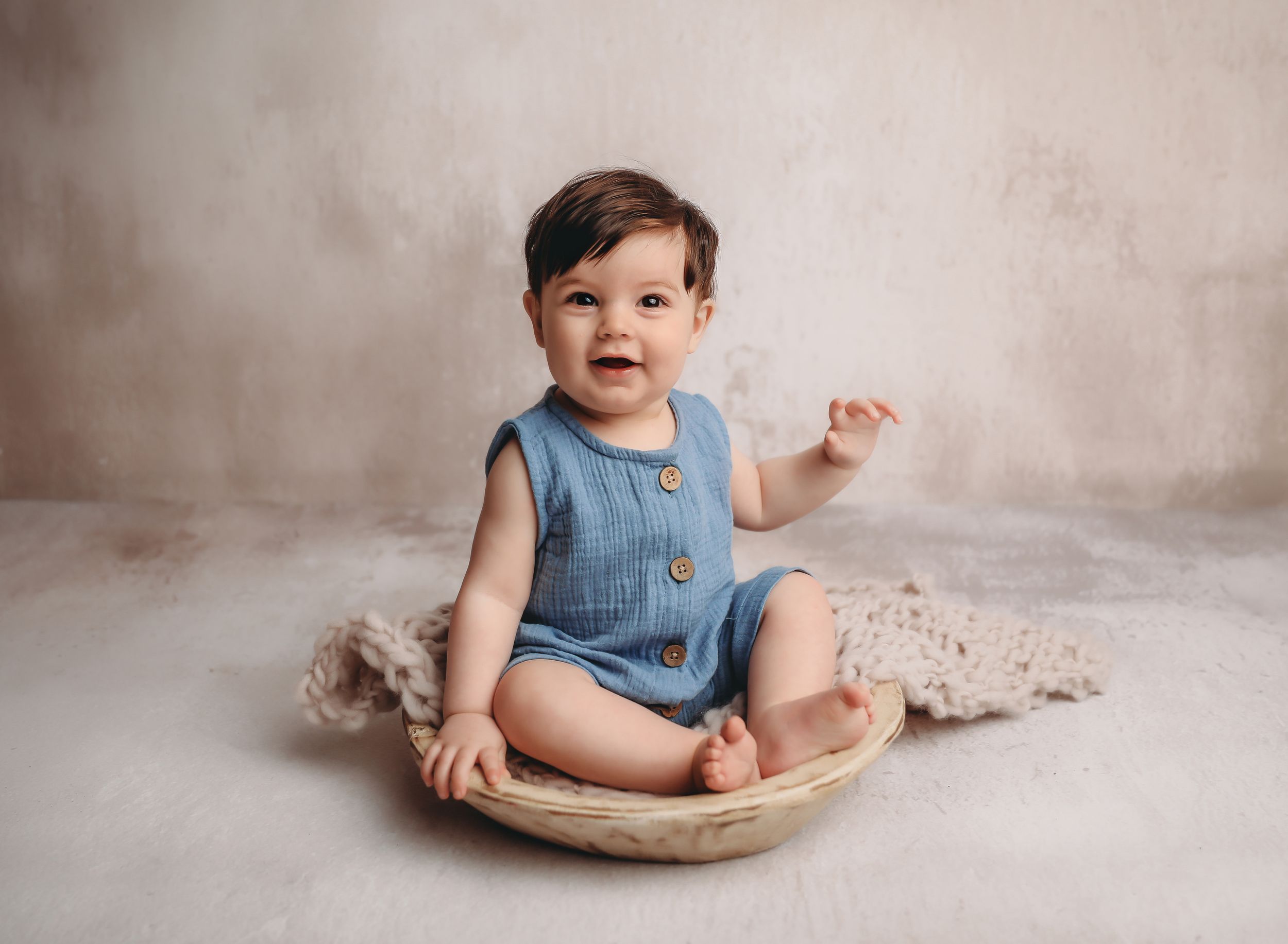 Smiling baby in blue romper sitting in wooden bowl on soft blanket against neutral backdrop.