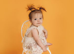 Adorable baby girl in lace dress with pigtails sitting on white chair against bright yellow background.