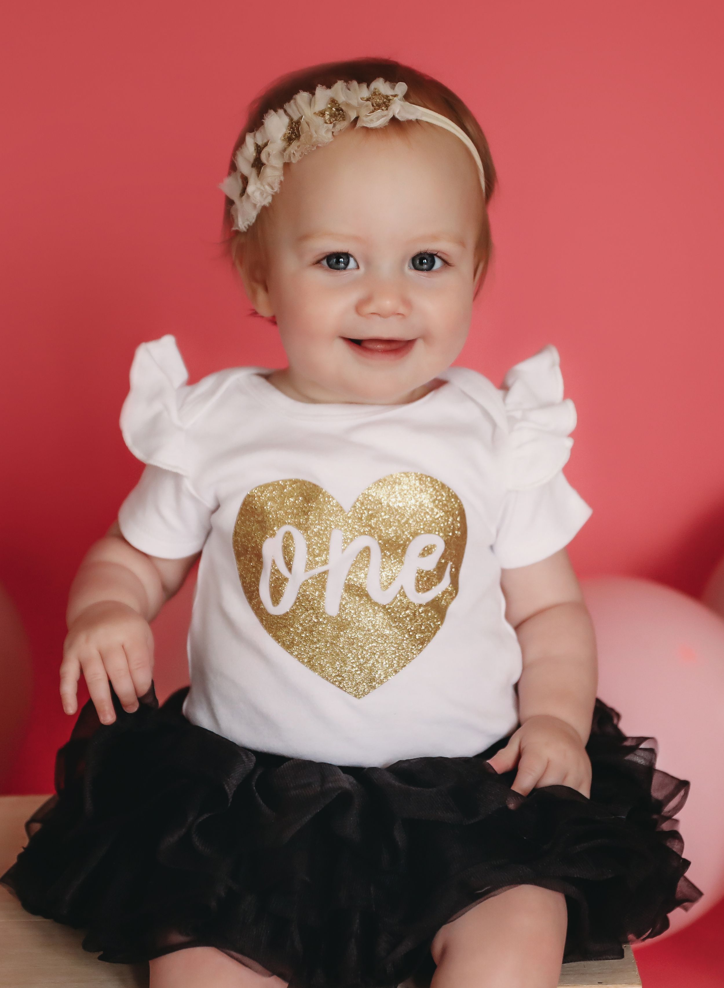 Baby girl in white “one” birthday onesie with gold glitter heart, black tutu skirt, and floral headband smiling at camera.