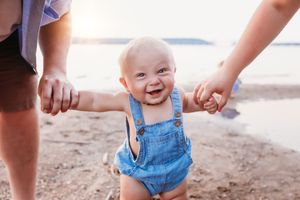 A toddler in denim overalls takes first steps on a sandy beach while being supported by adult hands.