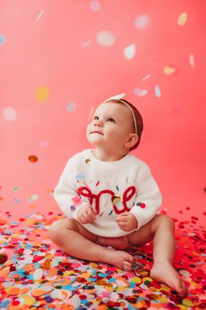 Baby in white onesie lying on floor surrounded by colorful confetti against coral pink background.