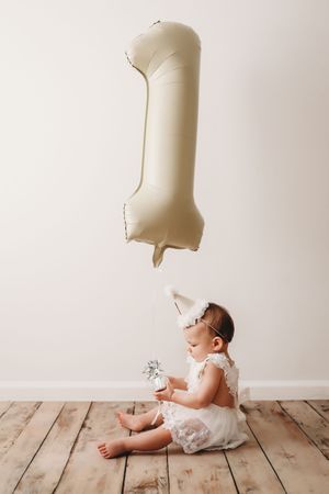A baby in white clothing plays with floating milk bottles in a minimalist photo series against a light background.