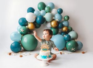 First birthday cake smash photo session with teal, gold, and white balloon arch decoration in the background.