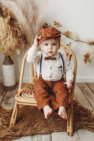 Baby in polka dot bib and brown overalls sits in rattan chair against neutral backdrop with dried florals.