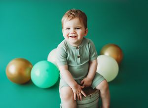 Baby in gray onesie sits on teal backdrop with gold and mint balloons for milestone photo session.