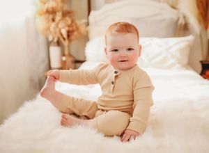 Baby wearing cozy beige onesie sits on white fluffy blanket in warm-toned nursery setting.